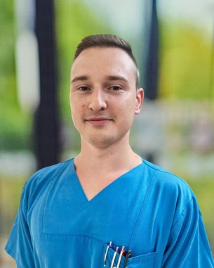 Ein junger Mann in einem blauen medizinischen Kittel steht mit einem neutralen Ausdruck. In seiner Brusttasche sind mehrere Stifte sichtbar.