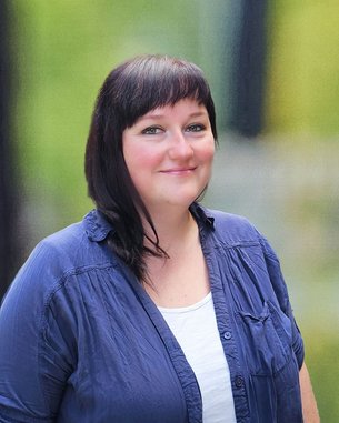 Eine Frau mit schulterlangen, dunklen Haaren trägt ein blaues Hemd und steht lächelnd vor einem unscharfen, grünen Hintergrund.