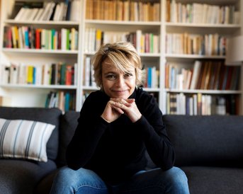 Eine Person mit kurzen, blonden Haaren sitzt auf einem grauen Sofa, die Hände unter dem Kinn verschränkt. Im Hintergrund sind Bücherregale mit einer Vielzahl von Büchern sichtbar.