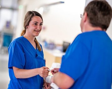 Eine Krankenschwester in blauer Arbeitskleidung spricht mit einem Kollegen. Beide stehen in einem Krankenhausflur. Die Krankenschwester hält einen Stift und scheint Informationen auszutauschen.