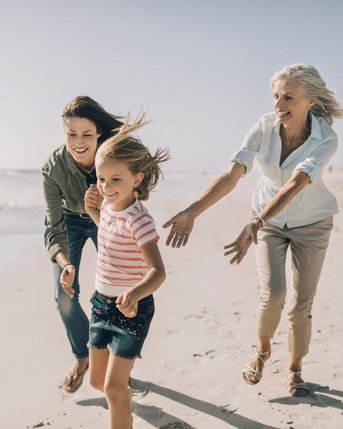 Drei Frauen, darunter ein Mädchen, laufen fröhlich am Strand entlang. Die Frauen tragen lässige Kleidung und scheinen Spaß zu haben, während sie dem Mädchen hinterherlaufen. Das Meer ist im Hintergrund sichtbar.