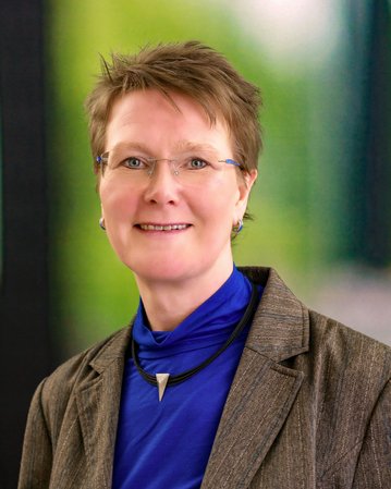 Eine Frau mit kurzen, blonden Haaren und Brille trägt einen grauen Blazer über einem blauen Oberteil. Der Hintergrund ist unscharf und grün.