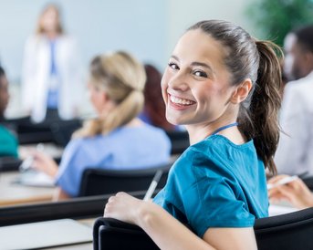 Eine junge Frau mit einem freundlichen Lächeln sitzt in einem Klassenzimmer, während sie sich umdreht. Im Hintergrund sind weitere Teilnehmer in medizinischer Kleidung zu sehen, die auf einen Vortrag achten.