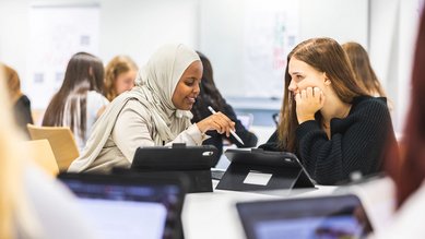 Zwei Schülerinnen sitzen an einem Tisch und diskutieren, während sie Tablets benutzen. Im Hintergrund sind weitere Schüler zu sehen.