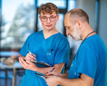 Zwei Personen in blauen medizinischen Kitteln stehen in einem modernen Raum. Eine Person, ein junger Mann mit lockigem Haar und Brille, hält ein Notizbuch und einen Stift, während die andere Person, ein älterer Mann, Notizen macht.