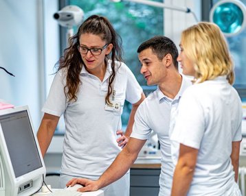 Drei medizinische Fachkräfte in weißen Kitteln stehen um einen Computer. Sie betrachten den Bildschirm und diskutieren Informationen. Im Hintergrund sind medizinische Geräte und ein Fenster sichtbar.