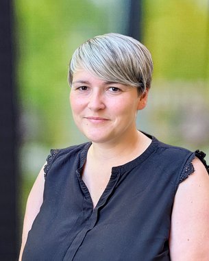 Eine Frau mit kurzem, grauem Haar trägt ein schwarzes Oberteil und steht vor einer unscharfen, hellen Hintergrundszene.