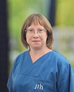 Eine Person in blauer Arbeitskleidung steht vor einem unscharfen Hintergrund. Sie trägt eine Brille und hat schulterlange Haare.