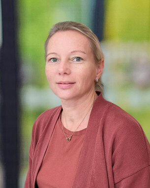 Eine Frau mit blonden Haaren trägt einen roten Cardigan und schaut direkt in die Kamera. Der Hintergrund ist unscharf und grün.
