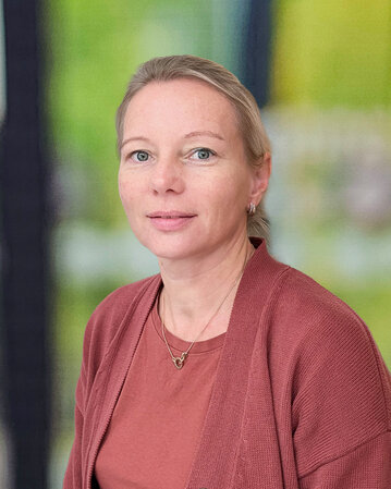 Eine Frau mit blonden Haaren trägt einen roten Cardigan und schaut direkt in die Kamera. Der Hintergrund ist unscharf und grün.