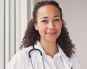 Eine Ärztin mit lockigem Haar trägt einen weißen Kittel und ein Stethoskop um den Hals. Sie steht in einem hellen Raum mit großen Fenstern und lächelt freundlich.