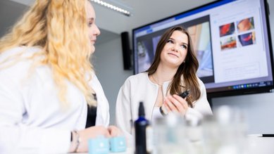 Zwei Frauen in weißen Kitteln sitzen an einem Tisch, während eine von ihnen ein Produkt in der Hand hält. Im Hintergrund ist ein Bildschirm mit Bildern zu sehen.