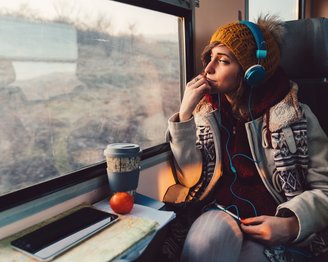 Eine Frau mit Kopfhörern sitzt in einem Zug und schaut aus dem Fenster. Sie trägt eine Mütze und einen dicken Mantel. Auf dem Tisch vor ihr liegen ein Smartphone, ein Notizbuch und eine orangefarbene Frucht.