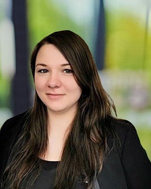 Eine Person mit langen, dunklen Haaren und einem schwarzen Oberteil, die in einem hellen, unscharfen Hintergrund steht.