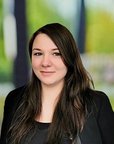 Eine Person mit langen, dunklen Haaren und einem schwarzen Oberteil, die in einem hellen, unscharfen Hintergrund steht.