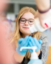 Eine Person mit Brille und langen Haaren trägt blaue Handschuhe und hält eine Handdesinfektionsflasche, während jemand anderes sie anreicht.