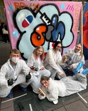 Fünf Personen in Schutzanzügen posieren vor einem bunten Graffiti mit dem Schriftzug "SIM 2025" und halten Spraydosen in der Hand.
