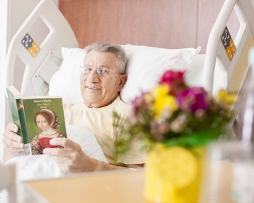 Ein älterer Mann im Krankenhausbett liest ein Buch. Er trägt eine Brille und lächelt. Auf dem Nachttisch steht eine blühende Pflanze in einem gelben Topf.