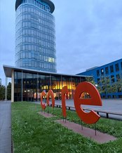 Moderne Glasgebäude mit einem großen roten Schriftzug "core" im Vordergrund, umgeben von Rasenflächen und Bänken.