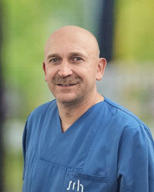 Ein Mann mit Glatze trägt ein blaues medizinisches Oberteil und lächelt. Im Hintergrund sind unscharfe grüne und graue Elemente sichtbar.