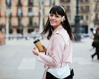 Eine Frau mit Kopfhörern trägt eine rosa Lederjacke und hält einen Kaffeebecher in der Hand. Sie lächelt und blickt über die Schulter, während Passanten im Hintergrund vorbeigehen.