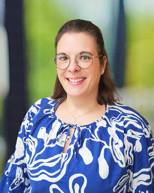 Eine lächelnde Frau mit Brille trägt ein blaues, gemustertes Oberteil. Der Hintergrund zeigt unscharfe grüne und helle Elemente.