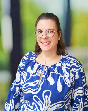 Eine lächelnde Frau mit Brille trägt ein blaues, gemustertes Oberteil. Der Hintergrund zeigt unscharfe grüne und helle Elemente.