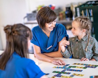 Eine Erwachsene in blauer Kleidung interagiert mit einem kleinen Mädchen, das eine grüne Bluse trägt. Beide sitzen an einem Tisch, auf dem bunte Spielkarten verteilt sind. Eine weitere Person ist im Hintergrund sichtbar.