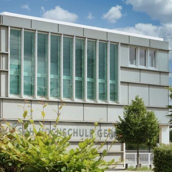 Moderne Schulgebäude mit großen Fenstern und einer klaren, geometrischen Fassade. Der Schriftzug "Hochschule" ist an der Vorderseite sichtbar, umgeben von Bäumen und Sträuchern.