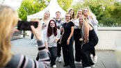 Gruppe von neun Personen posiert für ein Foto im Freien, während eine Person im Vordergrund das Bild mit einem Smartphone aufnimmt.