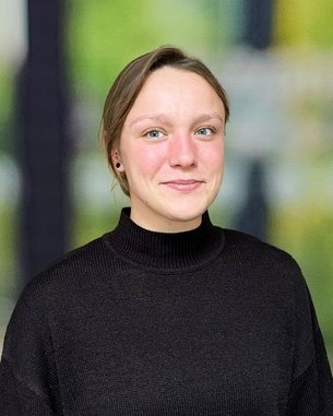 Eine Person mit kurzen Haaren trägt einen schwarzen Pullover und steht vor einem unscharfen, grünen Hintergrund.