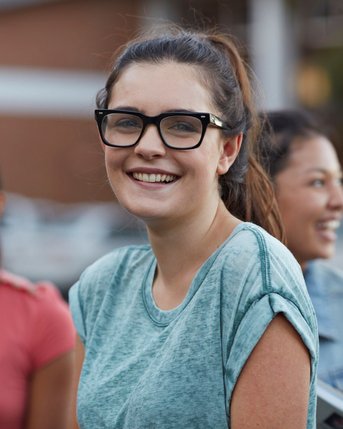 Eine Gruppe von vier jungen Erwachsenen steht zusammen, während eine Frau mit Brille in der Mitte lächelt. Sie trägt ein graues T-Shirt und hat lange, lockige Haare.