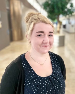 Eine junge Frau mit blonden Haaren und einem gepunkteten Oberteil steht in einem modernen Innenraum mit Pflanzen im Hintergrund.