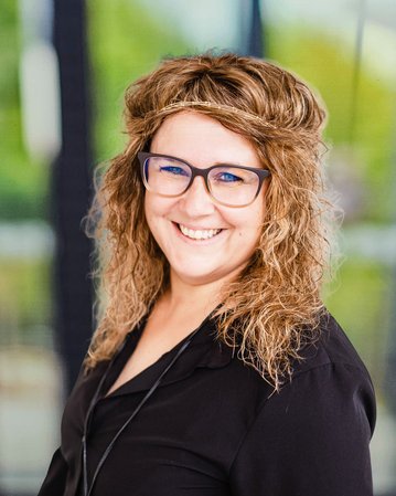 Frau mit lockigem, braunem Haar und Brille, trägt ein schwarzes Oberteil und lächelt freundlich in die Kamera.