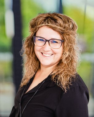 Frau mit lockigem, braunem Haar und Brille, trägt ein schwarzes Oberteil und lächelt freundlich in die Kamera.