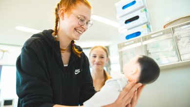 Eine Schülerin übt mit einer Puppenpuppe, während eine andere Schülerin im Hintergrund zuschaut. Beide sind in einem Klassenzimmer.