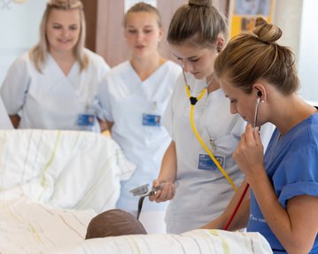 Eine Gruppe von Pflegekräften in weißen Uniformen und einer blauen Uniform steht um ein Bett. Eine Krankenschwester verwendet ein Stethoskop, während eine andere eine Messung vornimmt.