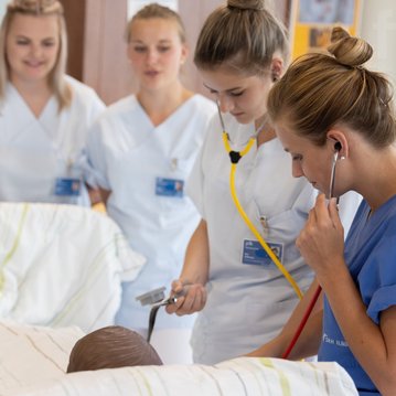 Eine Gruppe von Pflegekräften in weißen Uniformen und einer blauen Uniform steht um ein Bett. Eine Krankenschwester verwendet ein Stethoskop, während eine andere eine Messung vornimmt.