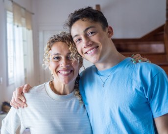 Eine Frau mit lockigem Haar und ein junger Mann mit kurzem Haar stehen lächelnd nebeneinander in einem hellen Raum.