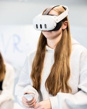 Eine Person mit langen Haaren trägt ein VR-Headset und hält einen Controller in der Hand, während sie in einem modernen Raum steht.