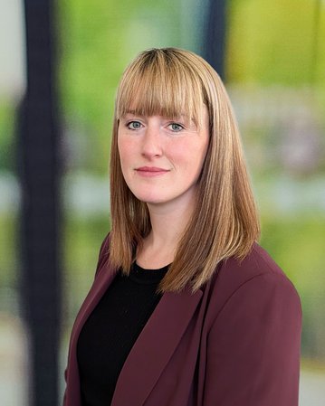 Eine Frau mit langen, blonden Haaren trägt einen burgunderfarbenen Blazer und steht vor einem unscharfen Hintergrund mit grünen und grauen Tönen.