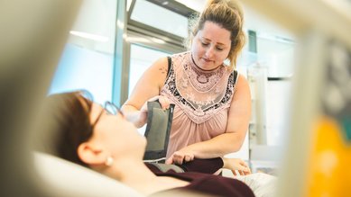 Eine Pflegekraft hilft einer Patientin, indem sie einen Verband anlegt. Die Patientin liegt auf einem Bett und trägt eine Brille.