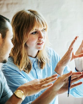 Eine Frau und ein Mann diskutieren vor einem Computerbildschirm. Die Frau gestikuliert mit den Händen, während sie spricht, und der Mann hört aufmerksam zu. Beide scheinen an einem kreativen oder beruflichen Projekt zu arbeiten.