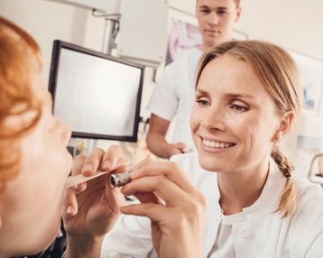 Eine Ärztin untersucht das Maul eines Patienten mit einem medizinischen Instrument, während ein weiterer Mitarbeiter im Hintergrund beobachtet. Beide tragen medizinische Kleidung in einer klinischen Umgebung.