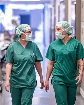 Zwei Krankenschwestern in grüner Arbeitskleidung und Mundschutz gehen durch einen Krankenhausflur. Im Hintergrund sind weitere medizinische Fachkräfte und Krankenhausausstattung sichtbar.
