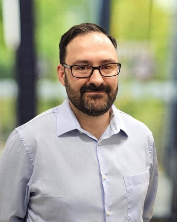 Ein Mann mit Brille und Bart trägt ein hellblaues Hemd und steht vor einem unscharfen, grünen Hintergrund.