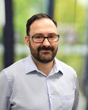 Ein Mann mit Brille und Bart trägt ein hellblaues Hemd und steht vor einem unscharfen, grünen Hintergrund.