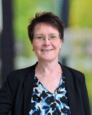 Eine lächelnde Frau mit kurzen, braunen Haaren trägt eine blumige Bluse und einen schwarzen Blazer. Sie steht vor einem unscharfen Hintergrund.