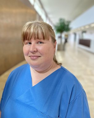 Eine Person mit blauer Arbeitskleidung steht in einem modernen Innenraum mit Holz- und Pflanzenelementen im Hintergrund.