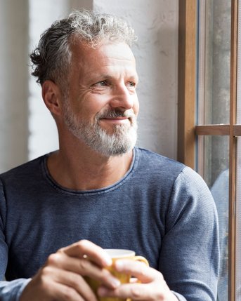 Ein lächelnder Mann mit grauem Bart hält eine Tasse in der Hand und schaut nachdenklich aus einem Fenster.